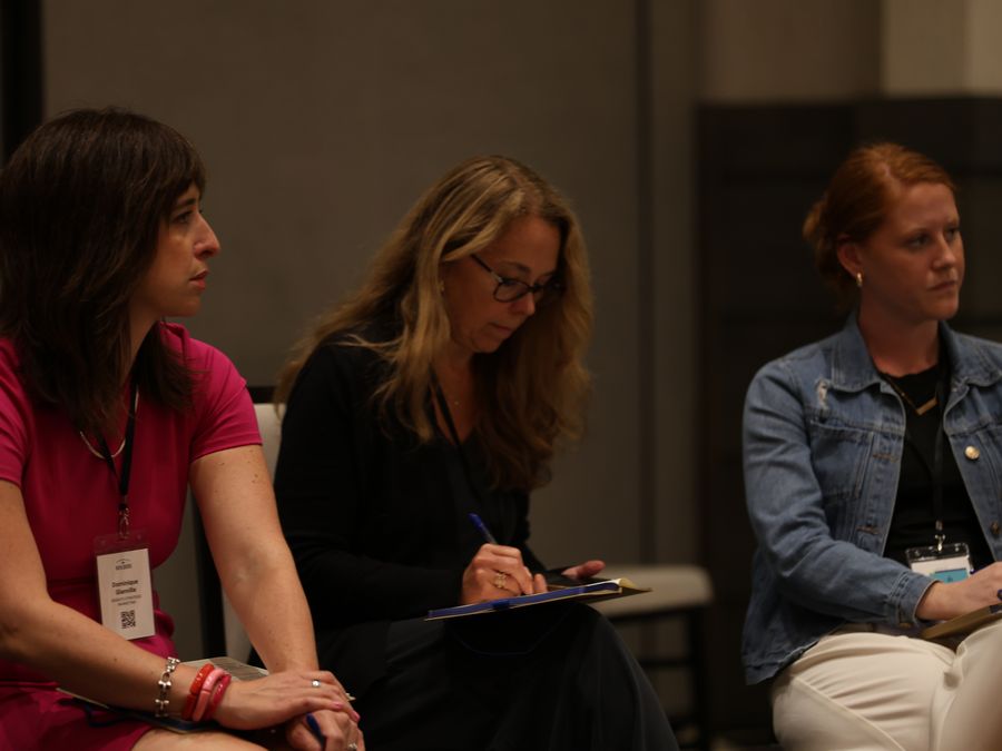 Three attendees listen quietly, one writing notes on a clipboard